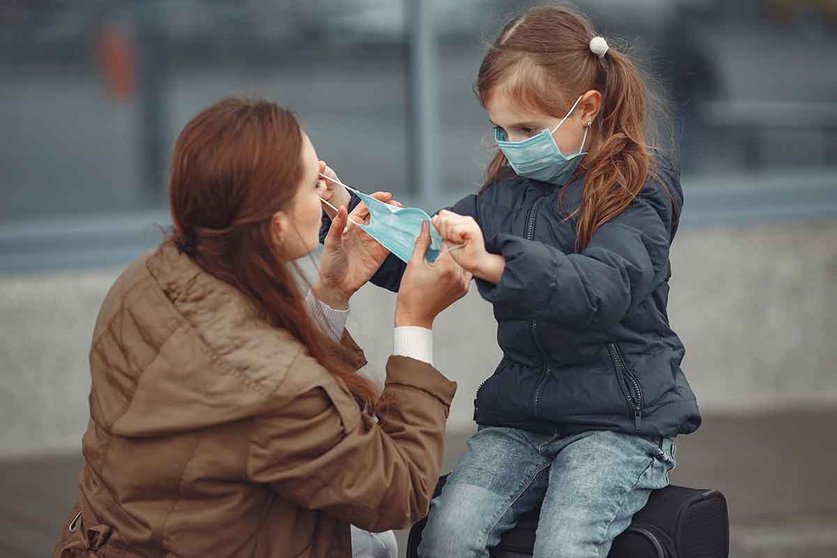 A European mother in a respirator with her daughter are standing near a building.The parent is teaching her child how to wear protective mask to save herself from virus.