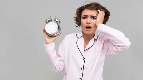 Portrait of a young confused girl in pajamas holding alarm clock and looking at camera isolated over gray background